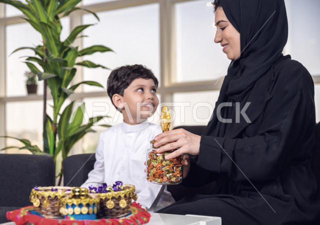 A wonderful participation of a Saudi Arabian Gulf mother with her son on the morning of Eid, offering him sweets with gestures indicating happiness and joy, wearing traditional Saudi dress, Eidiya, Gergean, celebrations and manifestations of Eid, spending