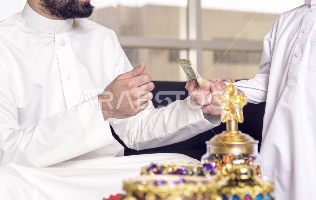 A wonderful participation of a Saudi Arabian Gulf father with his son on the morning of Eid, presenting the Eid with happiness, wearing traditional Saudi dress, Eidiya, Gergean, celebrations and manifestations of Eid, an official holiday, spending happy f