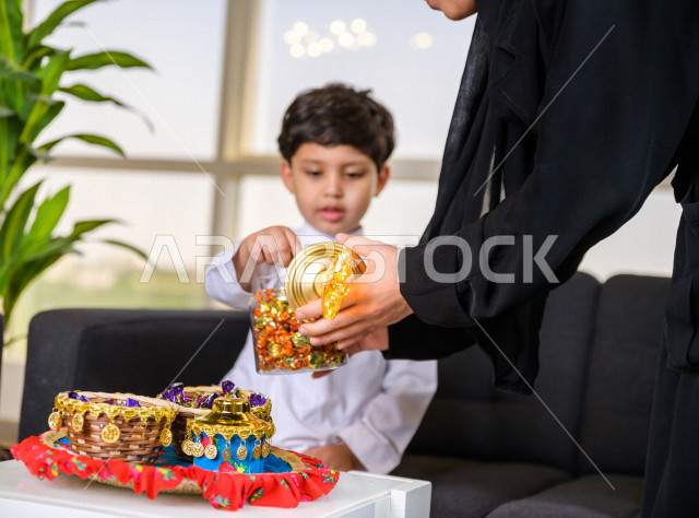 A wonderful participation of a Saudi Arabian Gulf mother with her son on the morning of Eid, offering him sweets with gestures indicating happiness and joy, wearing traditional Saudi dress, Eidiya, Gergean, celebrations and manifestations of Eid, spending
