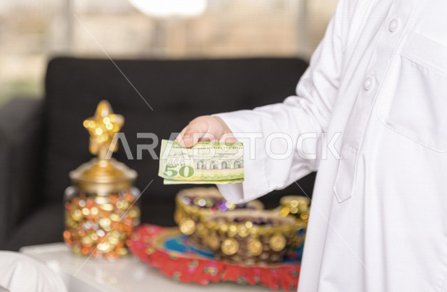 صورة مقربة لطفل سعودي خليجي في صباح يوم العيد يمسك بيده عيدية العيد ، يرتدي اللباس السعودي التقليدي ، العيدية ، القرقيعان ، احتفالات و مظاهر العيد ، قضاء أوقات عائلية سعيدة في العيد