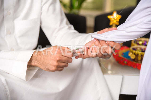 صورة مقربة لمشاركة رائعة لأب عربي خليجي سعودي مع ابنه في صباح يوم العيد ، تقديم العيدية بسعادة ، ارتداء اللباس السعودي التقليدي ، العيدية ، القرقيعان ، احتفالات و مظاهر العيد ، عطلة رسمية ، قضاء أوقات عائلية سعيدة في العيد