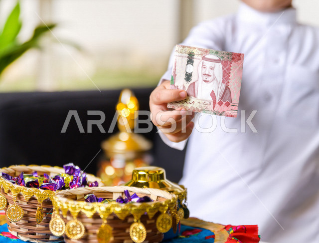 صورة مقربة لطفل سعودي خليجي في صباح يوم العيد يمسك بيده عيدية العيد ، يرتدي اللباس السعودي التقليدي ، العيدية ، القرقيعان ، احتفالات و مظاهر العيد ، قضاء أوقات عائلية سعيدة في العيد
