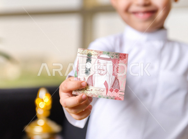 صورة مقربة لطفل سعودي خليجي في صباح يوم العيد يمسك بيده عيدية العيد بسعادة ، يرتدي اللباس السعودي التقليدي ، العيدية ، القرقيعان ، احتفالات و مظاهر العيد ، قضاء أوقات عائلية سعيدة في العيد