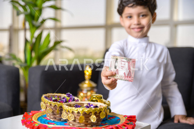 طفل سعودي خليجي في صباح يوم العيد إيماءات و تعابير وجه تدل على السعادة بأخذ عيدية العيد  ، يرتدي اللباس السعودي التقليدي ، العيدية ، القرقيعان ، احتفالات و مظاهر العيد ، قضاء أوقات عائلية سعيدة في العيد