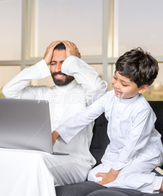 رجل اعمال عربي سعودي خليجي يعمل عن بعد في المنزل لضبط انتشار العدوى ، استخدام الكمبيوتر المحمول لانجاز العمل ، يشعر بالانزعاج بسبب تشويش الابن له اثناء العمل ، اجتماعات اونلاين