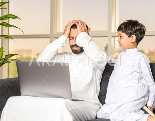 رجل اعمال عربي سعودي خليجي يعمل عن بعد في المنزل لضبط انتشار العدوى ، استخدام الكمبيوتر المحمول لانجاز العمل ، يشعر بالانزعاج بسبب تشويش الابن له اثناء العمل ، اجتماعات اونلاين