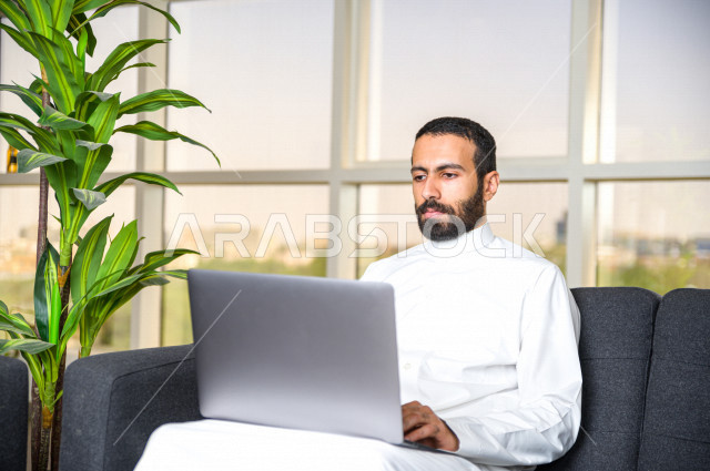 A Saudi Gulf businessman who works remotely at home to control the spread of infection, use a laptop computer to get the job done, feels happy to do the job successfully remotely, online meetings