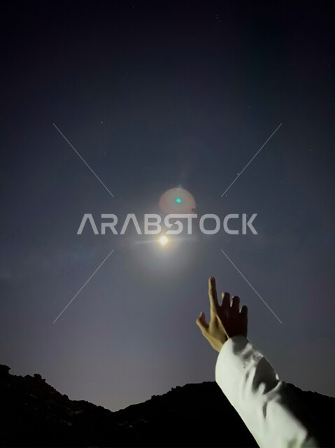 يد رجل يشير باصبع السبابة إلى القمر المضيء في السماء الصافية، ضوء القمر فوق ظل الجبال السوداء، سماء مدينة نجران 
