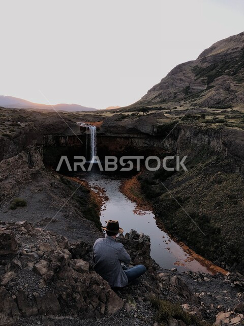 A man wearing a hat and sitting in front of a waterfall, a valley between mountains, mountains and rocky heights, natural scenery, quiet tourist places