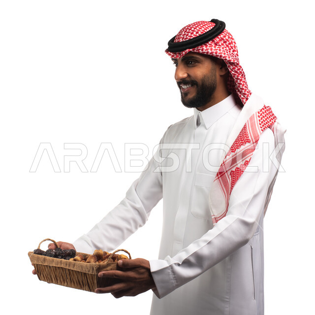 إيماءات وجه تدل على البشاشه والترحيب بالضيوف, بورتريه لرجل عربي سعودي خليجي يرتدي الزي السعودي التقليدي يحمل بيده سلة من القش مليئه بالتمر الطازج، حسن الضيافه في رمضان , منتج محلي صنع في السعودية ، منتجات محاصيل زراعية ، خلفيه بيضاء