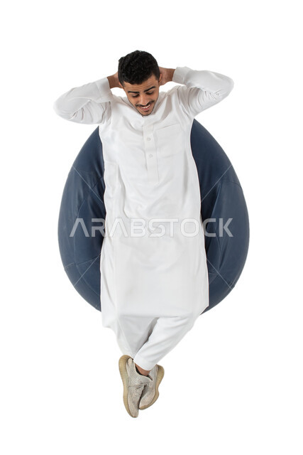 الحاجة إلى النوم ، ايماءات وجه ويدين تدل على الراحة والاسترخاء ، بورتريه علوي عامودي لشاب عربي سعودي خليجي يرتدي الثوب السعودي يجلس على كرسي طري ومريح، خلفية بيضاء