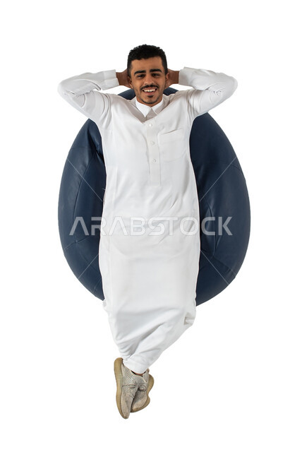 ايماءات وجه ويدين تدل على الراحة والاسترخاء ، بورتريه علوي عامودي لشاب عربي سعودي خليجي يرتدي الثوب السعودي يجلس على كرسي طري ومريح، خلفية بيضاء