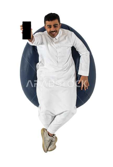 النظر إلى الكاميرا ، بورتريه علوي عامودي لرجل عربي سعودي خليجي يرتدي الثوب السعودي يجلس على كرسي,  تطبيقات وبرامج الجوال والتسوق الالكتروني , عرض شاشة سوداء فارغة ، خلفيه بيضاء
