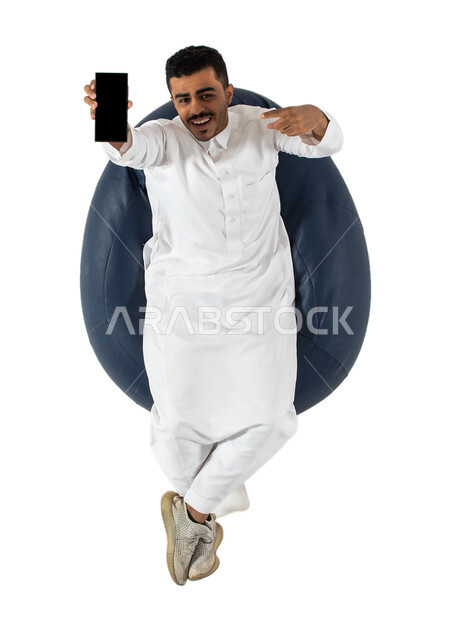 الاشاره الى الهاتف المحمول والنظر إلى الكاميرا ، بورتريه علوي عامودي لرجل عربي سعودي خليجي يرتدي الثوب السعودي يجلس على كرسي,  تطبيقات وبرامج الجوال والتسوق الالكتروني , عرض شاشة سوداء فارغة ، خلفيه بيضاء