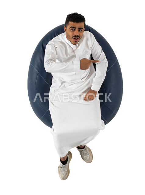 النظر إلى الكاميرا ، بورتريه علوي عامودي لرجل عربي سعودي خليجي يرتدي الثوب السعودي يجلس على كرسي, إيماءات يدين تشير الى شيء ما, حركات تدل على هذا المكان وهذا الموقع, خلفيه بيضاء