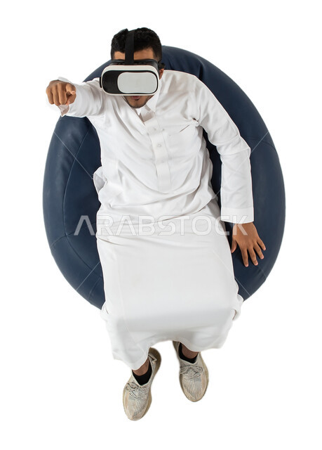 التفاعل والاندماج مع مشاهدة أفلام الواقع الافتراضي ، نظارة ثلاثية الابعاد، بورتريه علوي عامودي  لشاب عربي سعودي خليجي يرتدي الزي السعودي يجلس على كرسي مريح ويرتدي نظارة الواقع الافتراضي ،الاستمتاع بمشاهدة الأفلام وقضاء أوقات ممتعة ومسلية ، خلفية بيضاء