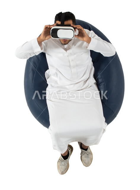 نظارة ثلاثية الابعاد، بورتريه علوي عامودي  لشاب عربي سعودي خليجي يرتدي الزي السعودي يجلس على كرسي مريح ويرتدي نظارة الواقع الافتراضي ،الاستمتاع بمشاهدة الأفلام وقضاء أوقات ممتعة ومسلية ، خلفية بيضاء