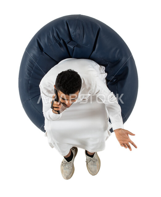 إتصال و تعارف وعلاقات اجتماعية ودردشة ومحادثات و تواصل اجتماعي  ،إجراء مكالمة هاتفية ،بورتريه علوي عامودي لرجل عربي سعودي خليجي مبتسم يرتدي الزي السعودي يجلس على كرسي مريح بيده هاتف محمول ،   خلفية بيضاء