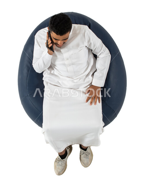 إجراء مكالمة هاتفية ،بورتريه علوي عامودي لرجل عربي سعودي خليجي مبتسم يرتدي الزي السعودي يجلس على كرسي مريح بيده هاتف محمول ،  إتصال و تعارف وعلاقات اجتماعية ودردشة ومحادثات و تواصل اجتماعي  ، خلفية بيضاء