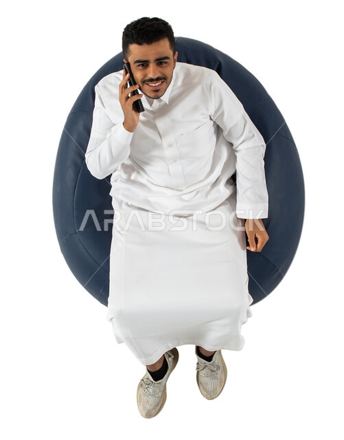 إجراء مكالمة هاتفية ،النظر إلى الكاميرا ، بورتريه علوي عامودي لرجل عربي سعودي خليجي مبتسم يرتدي الزي السعودي يجلس على كرسي مريح بيده هاتف محمول ،  إتصال و تعارف وعلاقات اجتماعية ودردشة ومحادثات و تواصل اجتماعي  ، خلفية بيضاء