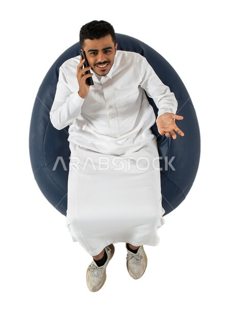 النظر إلى الكاميرا ، بورتريه علوي عامودي لرجل عربي سعودي خليجي مبتسم يرتدي الزي السعودي يجلس على كرسي مريح بيده هاتف محمول ، إجراء مكالمة هاتفية ، إتصال و تعارف وعلاقات اجتماعية ودردشة ومحادثات و تواصل اجتماعي  ، خلفية بيضاء