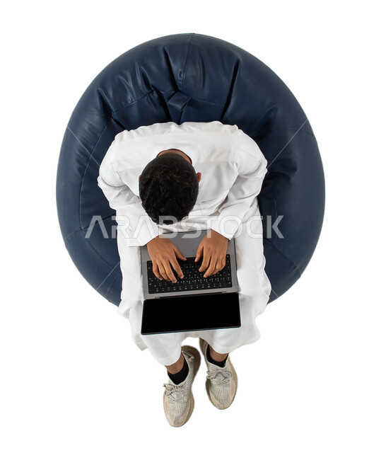 Chatting, conversations and social communication, online shopping and online requests, vertical portrait of a young Arab Saudi Gulf man wearing Saudi dress sitting on a comfortable chair, using a laptop, completing work tasks remotely, white background