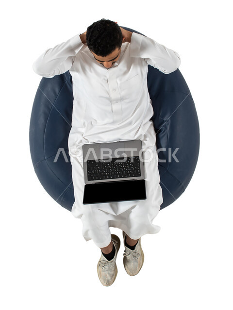 الاستمتاع بمشاهدة شي ما عبر جهاز الحاسوب المحمول ، دردشة ومحادثات وتواصل اجتماعي ، بورتريه علوي عامودي لرجل عربي سعودي خليجي يرتدي الزي السعودي يجلس على كرسي مريح  ،  بث مباشر ، خلفية بيضاء