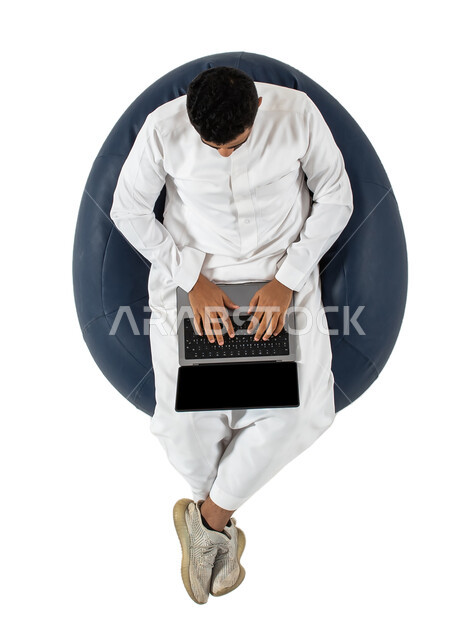 انجاز مهام العمل عن بعد, التسوق الالكتروني وطلبات الاونلاين ، بورتريه علوي عامودي  لرجل عربي سعودي خليجي يرتدي الزي السعودي يجلس على كرسي مريح ، العمل عن بعد باستخدام الحاسوب المحمول ،خلفية بيضاء