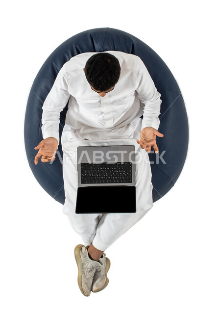 إيماءات يدين تدل على الدهشة والاستغراب ، بورتريه علوي عامودي لرجل عربي سعودي خليجي يرتدي الزي السعودي يجلس على كرسي مريح ، العمل عن بعد باستخدام الحاسوب المحمول ،خلفية بيضاء
