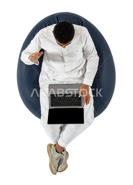 بث مباشر واجراء مكالمة فيديو عبر جهاز الكمبيوتر ،  بورتريه علوي عامودي لرجل عربي سعودي خليجي يرتدي الزي السعودي يجلس على كرسي مريح ، العمل عن بعد باستخدام الحاسوب المحمول ، خلفية بيضاء