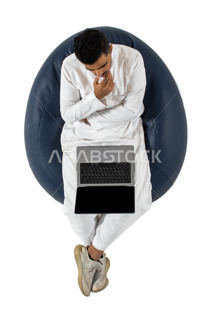 استخدام الحاسوب المحمول ، العمل عن بعد ، ايماءات يدين تدل على التفكير والتخطيط, حل المشكلات واتخاذ القرارات, بورتريه علوي عامودي لرجل عربي سعودي خليجي يرتدي الزي السعودي يجلس على كرسي مريح ، خلفية بيضاء