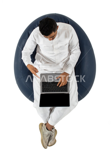 Live broadcast and video call via computer, working remotely using a laptop, vertical overhead portrait of a Saudi Arabian Gulf man wearing Saudi dress sitting on a comfortable chair, using a laptop, white background