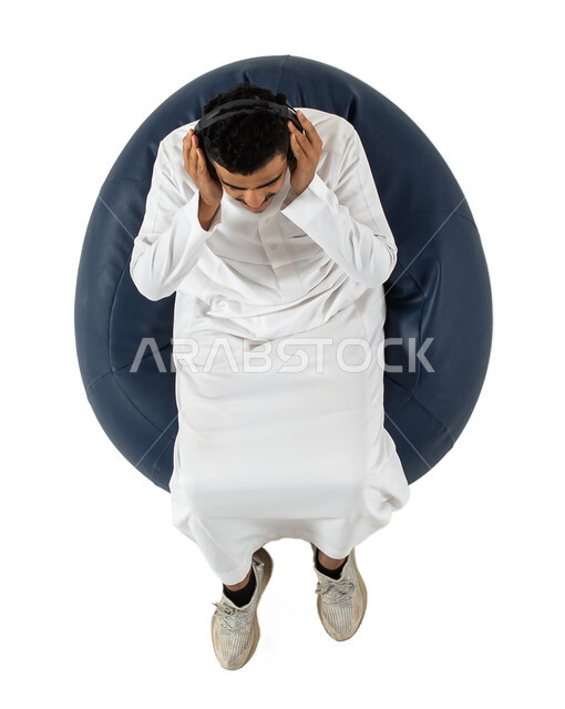 التسليه والترفيه باستخدام تقنية الهيدفون ، الاستمتاع بسماع الأغاني والموسيقى والصوتيات , بورتريه علوي عامودي  لرجل عربي سعودي خليجي يرتدي الزي السعودي يجلس على كرسي مريح يرتدي سماعة الرأس ،  خلفية بيضاء