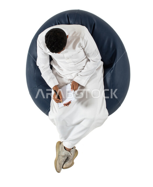 التمعن والتركيز ، إيماءات وجه ويدين تدل على التفكير و الحيرة ، كتابة وتسجيل الملاحظات ، بورتريه علوي عامودي  لرجل عربي سعودي خليجي يرتدي الزي السعودي يجلس على كرسي مريح ويمسك بيديه ورقة، حل المشكلات واتخاذ القرارات, خلفيه بيضاء