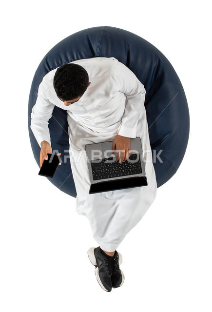 بث فيديو مباشر، اجتماع عمل عن بعد، بورتريه علوي عامودي لرجل عربي سعودي خليجي يرتدي الزي السعودي يجلس على كرسي مريح ، استخدام الهاتف المحمول والحاسوب المحمول، تواصل اجتماعي، محادثات ودردشة، خلفية بيضاء