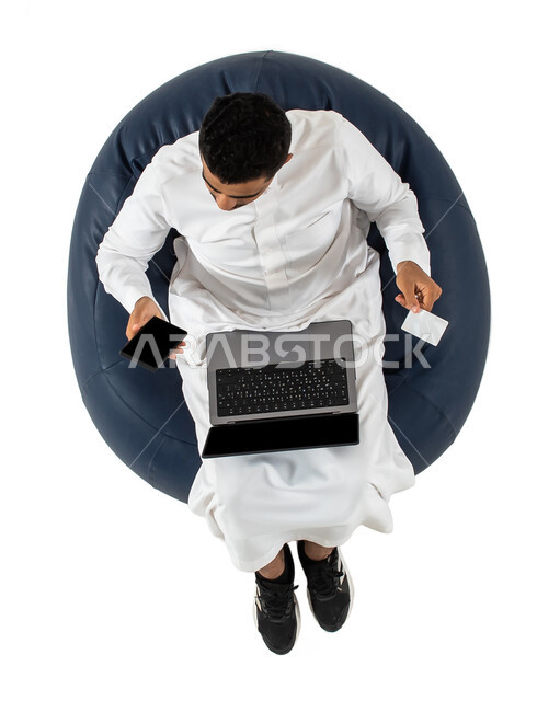 الدفع عن طريق البطاقه البنكيه, استخدام جهاز الحاسوب المحمول والهاتف المحمول للحجز الالكتروني ،  بورتريه علوي عامودي لشاب عربي سعودي خليجي يرتدي الزي السعودي يجلس على كرسي مريح ، الدفع الالكتروني ، خلفيه بيضاء