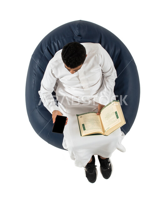 Spending time reading, learning and culture. A vertical portrait of a Saudi Arab man from the Gulf, dressed in Saudi dress, sitting on a comfortable chair, holding a book in his hands, and using a mobile phone, white background
