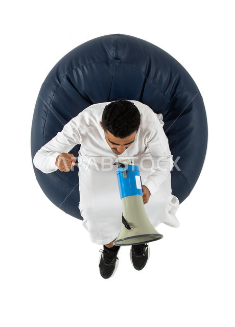  استخدام بوق الصوت ، ايماءات يد تدل على الهتاف والتشجيع والحماس, مشاهدة شيء ما ، بورتريه علوي عامودي لشاب عربي سعودي خليجي يرتدي الزي السعودي يجلس على كرسي ، يمسك بيده مكبر صوت للمناداه والاعلان ولفت الانتباه ، خلفية بيضاء