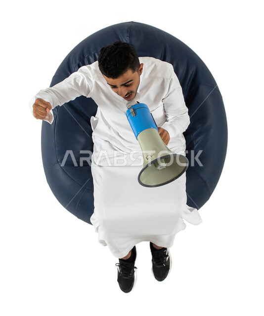 ايماءات يد تدل على الهتاف والتشجيع والحماس, مشاهدة شيء ما ، استخدام بوق الصوت ، بورتريه علوي عامودي لشاب عربي سعودي خليجي يرتدي الزي السعودي يجلس على كرسي ، يمسك بيده مكبر صوت للمناداه والاعلان ولفت الانتباه ، خلفية بيضاء