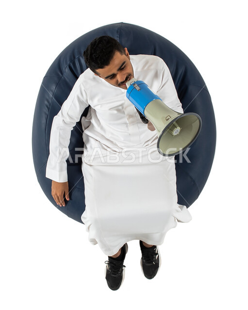 استخدام بوق الصوت ، بورتريه علوي عامودي لشاب عربي سعودي خليجي يرتدي الزي السعودي يجلس على كرسي ، يمسك بيده مكبر صوت للمناداه والاعلان ولفت الانتباه ،  خلفية بيضاء
