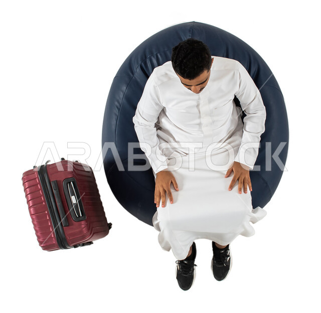Preparing for an enjoyable travel trip, a vertical overhead portrait of a young Saudi Arabian Gulf man wearing the Saudi dress, sitting on a chair with a travel bag next to him, waiting to board the plane, tourism and travel, white background