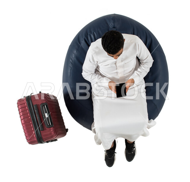 انتظار ركوب الطائره ، بورتريه علوي عامودي لرجل عربي سعودي خليجي يرتدي الزي السعودي يجلس على كرسي وبجانبه حقيبة سفر ، استخدام الهاتف المحمول ،  الاستعداد لرحلة سفر ممتعه ، سياحه وسفر ، خلفيه بيضاء