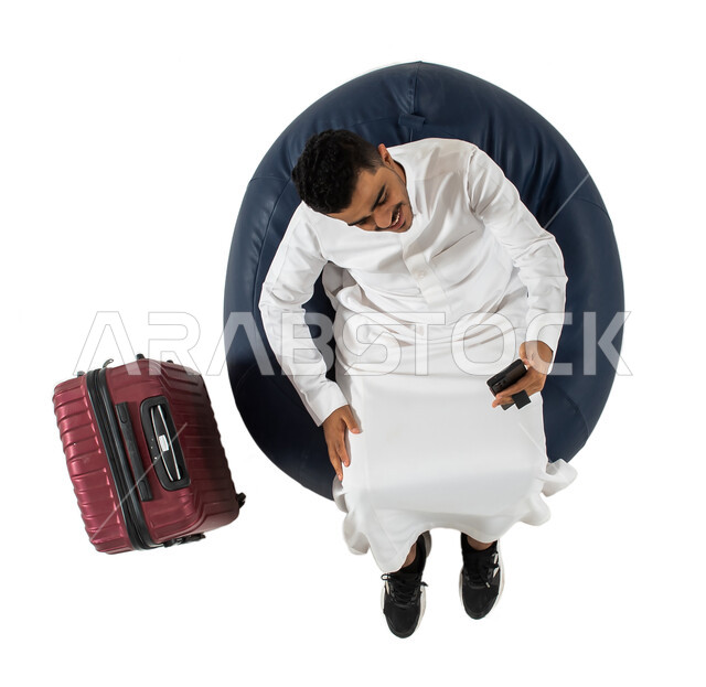 بث مباشر , اجراء مكالمة فيديو عبر الهاتف المحمول, بورتريه علوي عامودي لرجل عربي سعودي خليجي يرتدي الزي السعودي يجلس على كرسي وبجانبه حقيبة سفر ، استخدام الهاتف المحمول ، انتظار ركوب الطائره ، رحلة عمل ، سياحه وسفر ، خلفيه بيضاء