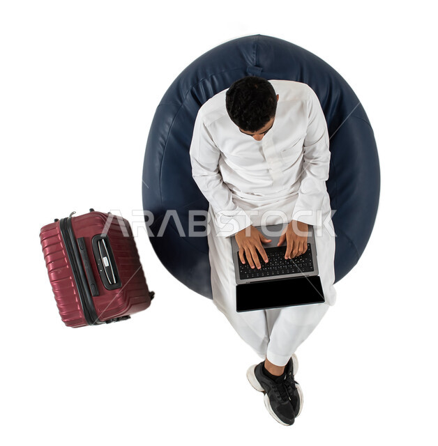 استخدام الحاسوب المحمول ، انجاز مهام العمل عن بعد ، بورتريه علوي عامودي لشاب عربي سعودي خليجي يرتدي الزي السعودي يجلس على كرسي وبجانبه حقيبة سفر ، انتظار ركوب الطائره ، رحلة عمل ، سياحه وسفر ، عطله ، خلفيه بيضاء