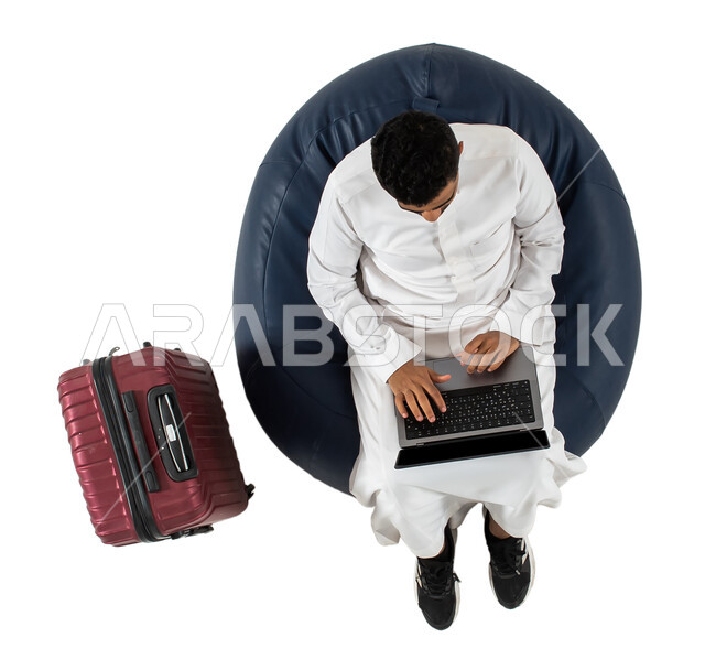 Completion of remote work tasks, a vertical overhead portrait of a Saudi Arab Gulf youth wearing the Saudi dress, sitting on a chair with a travel bag next to him, using a laptop, waiting to board a plane, business trip, tourism and travel, vacation, whit