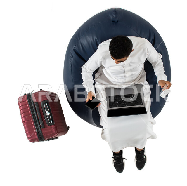 الدفع عن طريق البطاقه البنكيه, استخدام جهاز الحاسوب المحمول والهاتف المحمول للحجز الالكتروني ، انتظار ركوب الطائره  ، بورتريه علوي عامودي لشاب عربي سعودي خليجي يرتدي الزي السعودي يجلس على كرسي وبجانبه حقيبة سفر ، خلفيه بيضاء