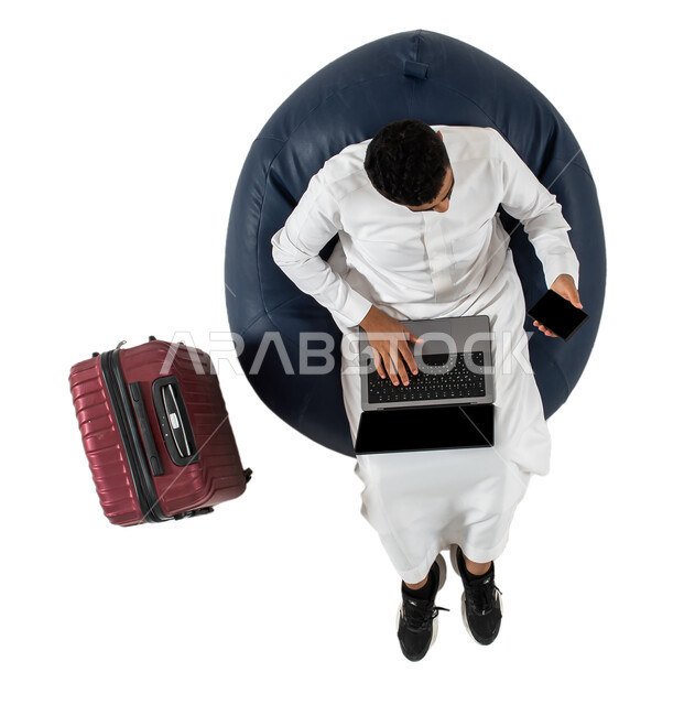استخدام الهاتف المحمول والحاسوب المحمول ، انتظار ركوب الطائره ، بورتريه علوي عامودي لرجل عربي سعودي خليجي يرتدي الزي السعودي يجلس على كرسي وبجانبه حقيبة سفر ، الاستعداد لرحلة سفر ممتعه ، رحلة عمل ، سياحه وسفر ، عطله صيفيه ، خلفيه بيضاء
