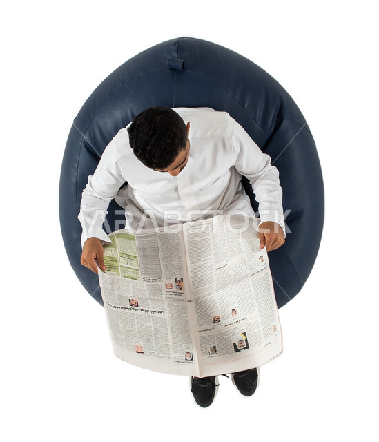 قضاء الوقت بالقراءه والتعلم والثقافة ، بورتريه علوي عامودي  لرجل عربي سعودي خليجي يرتدي الزي السعودي يجلس على كرسي مريح ويحمل بيديه جريدة ورقية ، تصفح الأخبار العالمية ، خلفية بيضاء