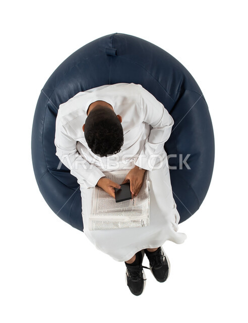 التسوق الإلكتروني، عرض خدمات وتطبيقات الهاتف المحمول، بورتريه علوي عامودي لرجل عربي سعودي خليجي يرتدي الزي السعودي يجلس على كرسي مريح، يمسك الهاتف المحمول , شاشة سوداء فارغة ،  استخدام جهاز الجوال ، خلفية بيضاء