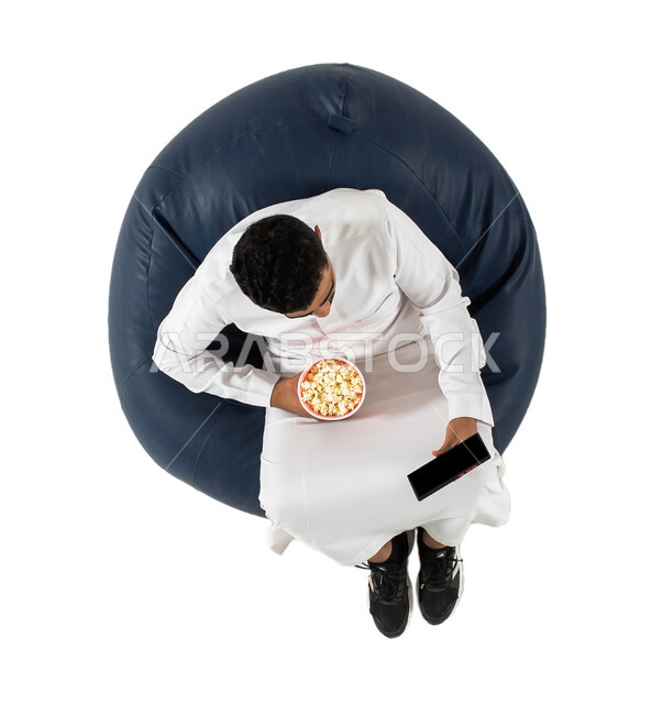 فيديو بث مباشر عبر الجوال, استخدام الهاتف المحمول ، بورتريه علوي عامودي لرجل عربي سعودي خليجي يرتدي الزي السعودي يجلس على كرسي يمسك بيده علبة الفوشار  , خلفية بيضاء